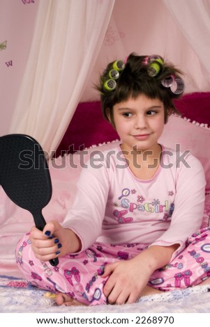 Adorable girl in pajamas with her hair in colorful curlers and looking in the mirror.