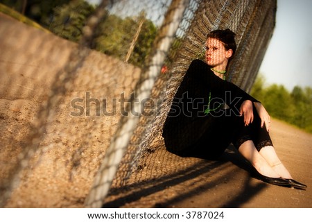 Attractive young lady sitting against a fence with a chain-link shadow criss-crossing her face.