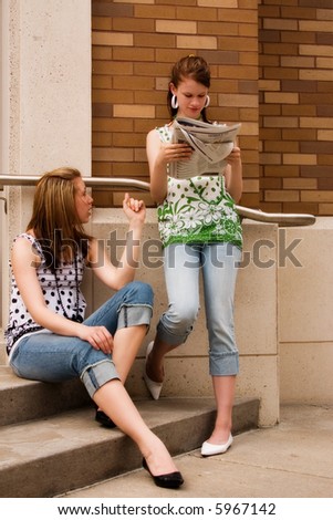 Two girls in the city, one leaning against a brick building while reading a newspaper, the other sitting on the stairs.