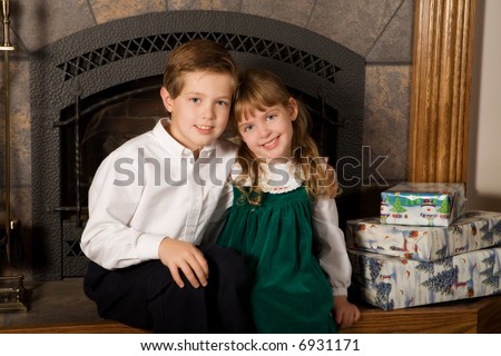 A young brother and sister sitting on the family hearth by wrapped Christmas gifts.