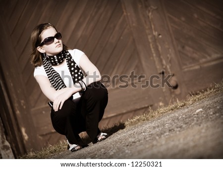 Preteen girl in sunglasses crouching outside by a barn door.  Sepia tone.