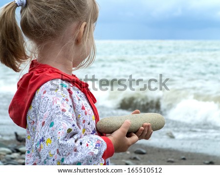 Little girl holding a big rock and turning back to look at storming sea