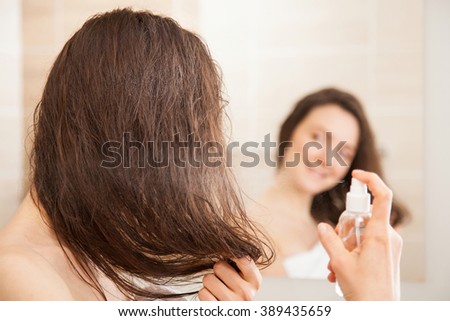 Smiling young woman applying hair spray in front of a mirror; haircare concept