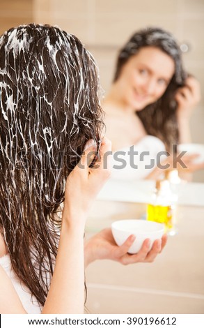 Smiling young woman applying hair mask in front of a mirror; haircare concept