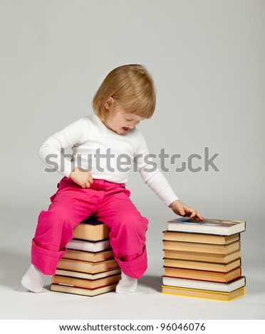 A funny little girl sitting on a stack of books and touching a book; neutral background