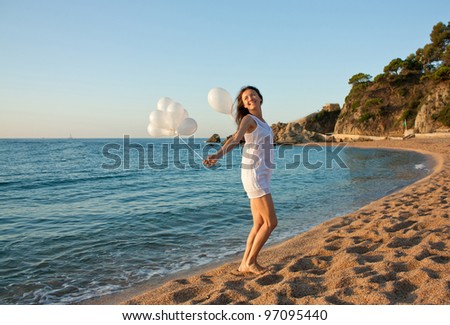 Happy smiling brunette girl with white balloons on sunny beach; attractive young woman enjoying summer vacation at the sea