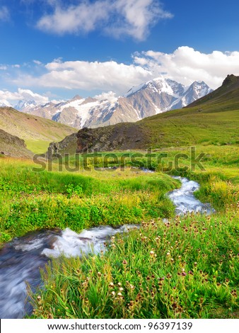 River on green meadow. High mountains and bright sky with clouds