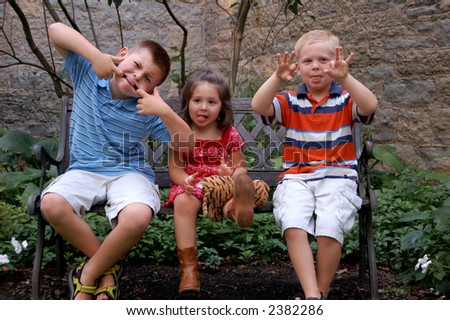 Three young children sitting on a bench making silly faces. Could be spring or summer.
