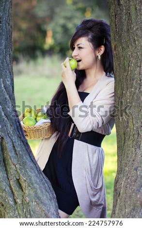 girl with a basket of apples in the hands, biting a green apple leaning on a tree