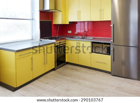 kitchen with yellow cupboards, topped with a grey worktops and fitted with appliances including refrigerator, microwave, oven, extractor hood, a wall with a red tile and floor with soft grey laminate