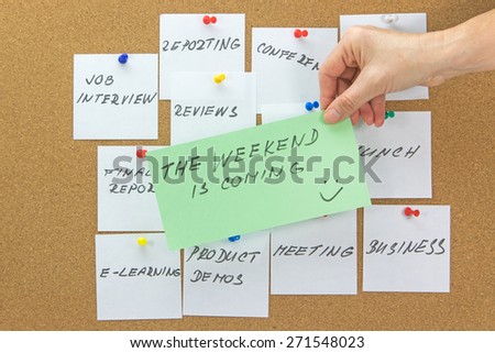 White paper notes with various written to-do tasks affixed to the corkboard.  Female hand holding a green card with the inscription \