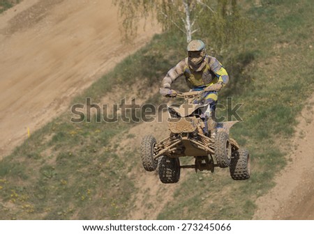 Quad racer is high jumping. The quad bike and rider are very muddy.Potential trademarks are removed and face of the racer is unidentifiable.