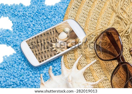 Straw hat, sunglasses and a seashell are lying on a little blue stones isolated on the white. Display of the white phone contains photo of seashells in basket. All potential trademarks are removed.