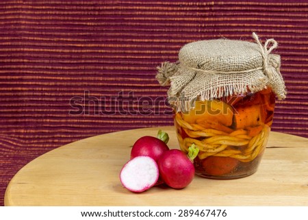 Jar of pickled variety of cheese in oil is standing on a wooden board. Reddish brown cloth background. Two whole and one halved radishes are lying next to the jar.