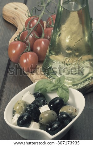 Vintage photo of white bowl with marinated green olives is placed on a wooden desk. Tomatoes and bottle of oil are lying on a desk of olive wood. Edited as a vintage photo. Vertically.