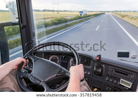 The driver is holding the steering wheel and is driving a truck on the highway. Empty road is ahead of him. The white truck is going in the opposite direction.