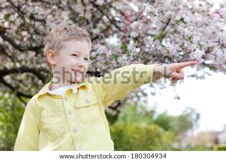 cheerful smiling little boy pointing at something with finger with beautiful blooming apple tree in the background at spring time