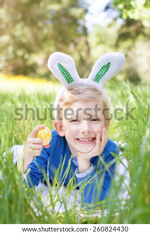little smiling boy with bunny ears holding easter egg after egg hunt in the park lying in the grass