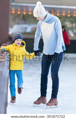 young mother teaching her positive son ice skating, enjoying winter time at outdoor skating rink together