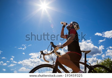 Cyclist resting and drinking isotonic drink. Backlight, sunny summer day.