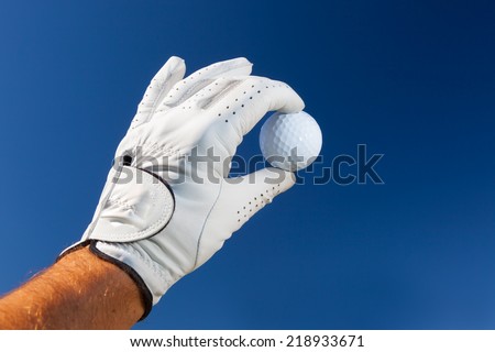 Hand wearing golf glove holding a white golf ball over a deep blue sky.