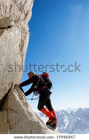 Climber climbing a rocky wall; vertical frame. Aiguille du Midi, Mont Blanc massif, Chamonix, France. In background the north face of Grand Jurasses peaks.