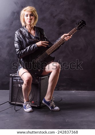 Beautiful young blonde siting on the amp dressed in black leather jacket with electric guitar on a black background