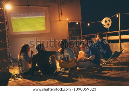 Group of young friends having fun while watching a football match on a building rooftop. Focus on the guy eating popcorn
