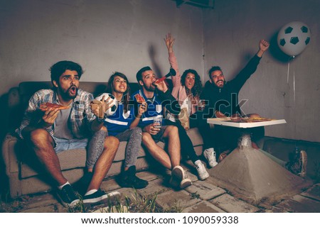 Group of young friends watching a football match on a building rooftop, cheering eating pizza and drinking beer, cheerful after they scored a goal