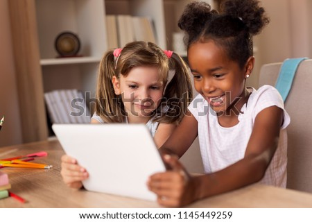 Two little girls sitting at a desk, playing video games on a tablet computer and having fun. Focus on the girl on the left
