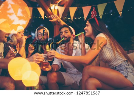 Group of young friends blowing party whistles, drinking beer and having fun at an outdoor New Year\'s Eve party. Focus on the guy on the right