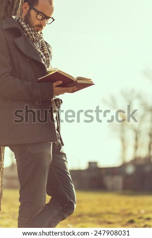 Man standing next to a tree and reading a book in the park on a sunny day