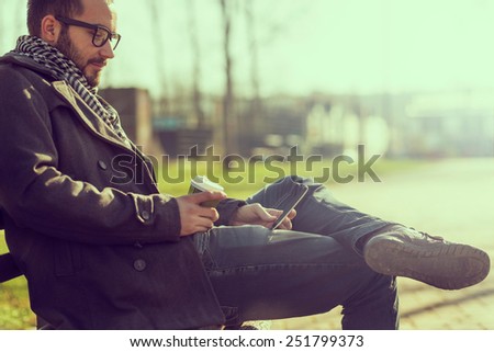 Young man sitting on a bench, reading an e-book on a tablet and drinking coffee