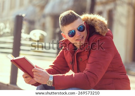 Young male sitting on a bench in an urban environment and reading a book