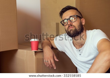 Young man moving in a new apartment, sitting on the floor, drinking take away coffee, surrounded with cardboard boxes