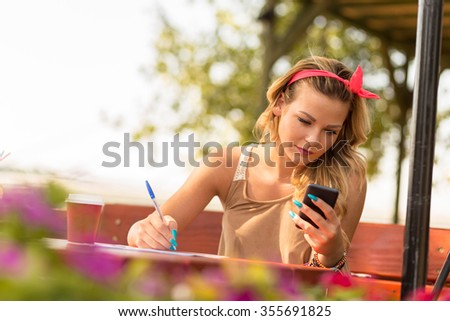 Beautiful attractive blond girl sitting in an outdoor cafe, holding a mobile phone and writing notes