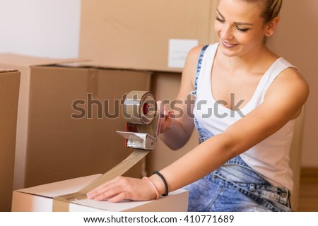 Young girl moving in a new apartment,standing surrounded with cardboard boxes, packing and taping boxes