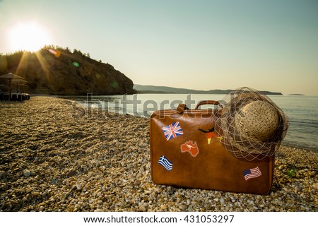 Vintage suitcase with flag stickers and a straw hat, placed on the beach with peaceful sea during sunrise on Thassos island in Greece