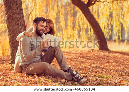 Couple in love sitting on autumn fallen leaves in a park, surfing the net on a tablet computer in search for travel destinations. Focus on a guy