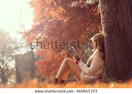 Beautiful young brunette sitting on a fallen autumn leaves in a park, reading a book