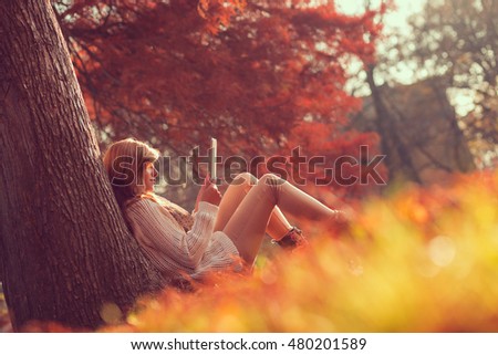 Beautiful young brunette sitting on a fallen autumn leaves in a park, reading an e-book on a tablet computer