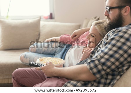 Young couple relaxing in their apartment, lying on the couch, watching a movie and eating popcorn. Lens flare effect on the window