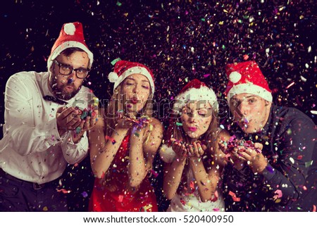Two beautiful young couples wearing Santa\'s hats, blowing colorful confetti at midnight at New Year\'s Eve party