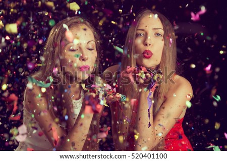 Two beautiful young women having fun at a Christmas party, blowing away confetti and sending kisses. Focus on the girl on the left