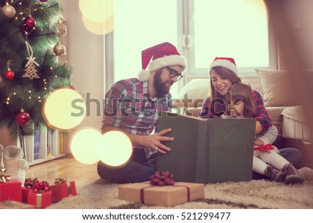 Young parents sitting on the living room floor next to a nicely decorated Christmas tree, mother holding a baby girl in her lap and all together reading a fairy tale