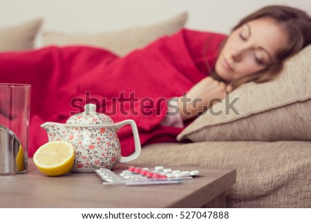 Sick woman covered with a blanket lying in bed with high fever and a flu, resting. Teapot, pills and lemon on the table, focus on the teapot