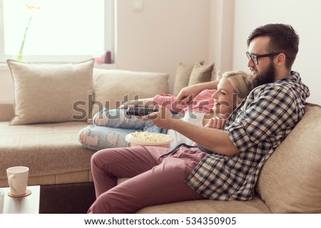 Young couple relaxing in their apartment, lying on the couch, watching a movie and eating popcorn. Lens flare effect on the window, focus on the guy