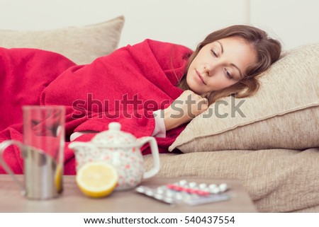 Sick woman covered with a blanket lying in bed with high fever and a flu, resting. Teapot, pills and lemon on the table