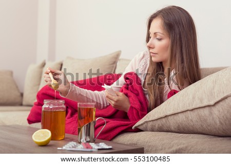 Sick woman lying in bed with high fever and a flu, putting some honey in her tea. Lemon, honey jar and pills set around the table