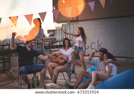 Young friends having fun at a rooftop party, playing the guitar, singing, dancing and chilling out. Focus on the girl playing the guitar